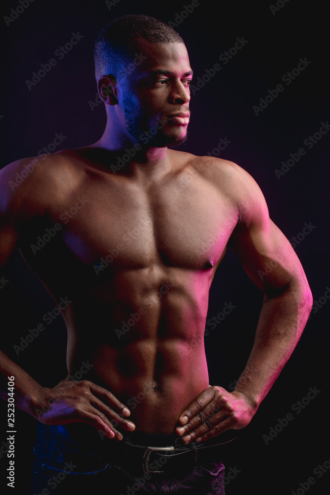African male athlete with naked torso with six pack posing against dark wall. Low key photo.