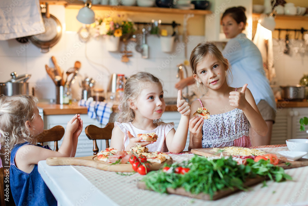 Kids are eating and tasting italian homemade pizza with mother. Cute children are enjoying delicious food in cozy home kitchen. Three girls at family dinner table. Lifestyle moment. Kids are eating and tasting italian homemade pizza with mother. Cute children are enjoying delicious food in cozy home kitchen. Three girls at family dinner table. Lifestyle moment.