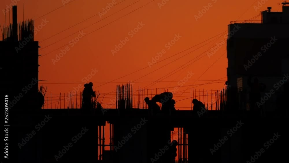 Construction of residential building. Builders go on the unfinished floor with protruding fittings. Silhouettes of workers on the background of an orange sunset. Construction of residential building. Builders go on the unfinished floor with protruding fittings. Silhouettes of workers on the background of an orange sunset.