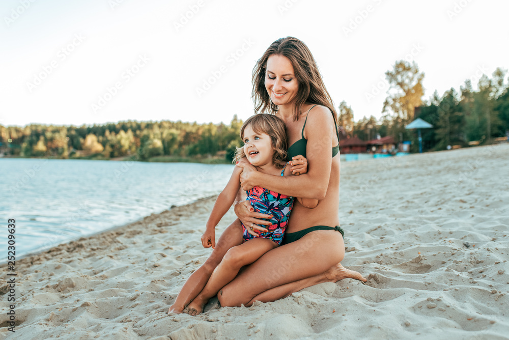 A young mother plays with her daughter girl of 2-5 years, on beach in summer, an outdoor resort by the lake. Happy rest on the weekend. Bright swimsuit on child. Parents love family and parenting. A young mother plays with her daughter girl of 2-5 years, on beach in summer, an outdoor resort by the lake. Happy rest on the weekend. Bright swimsuit on child. Parents love family and parenting.