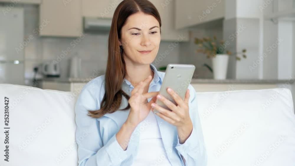 Young brunette woman with brown eyes sitting on sofa in living room using smartphone. Female housewife browsing on mobile phone chatting online with friends. Kitchen on background Young brunette woman with brown eyes sitting on sofa in living room using smartphone. Female housewife browsing on mobile phone chatting online with friends. Kitchen on background