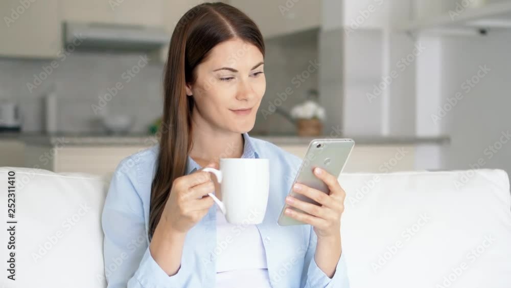 Brunette woman with brown eyes sitting in living room using smartphone. Young female browsing on mobile phone chatting online with friends. Housewife holding white coffee cup. Kitchen on background Brunette woman with brown eyes sitting in living room using smartphone. Young female browsing on mobile phone chatting online with friends. Housewife holding white coffee cup. Kitchen on background