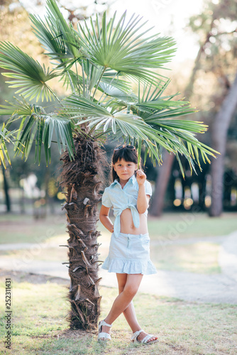 A pretty cute little girl in a beautiful summer suit stands by the palm trees