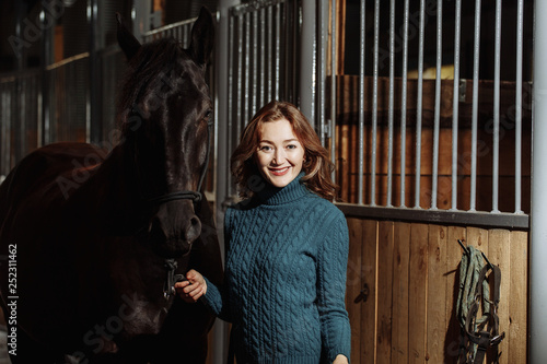 Young woman in sweater readying black horse for a ride while standing inside a stable on a farm