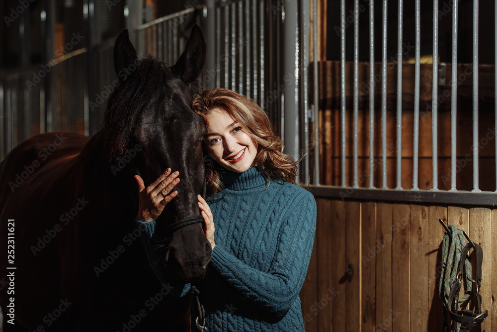 Young woman in sweater readying black horse for a ride while standing inside a stable on a farm Young woman in sweater readying black horse for a ride while standing inside a stable on a farm