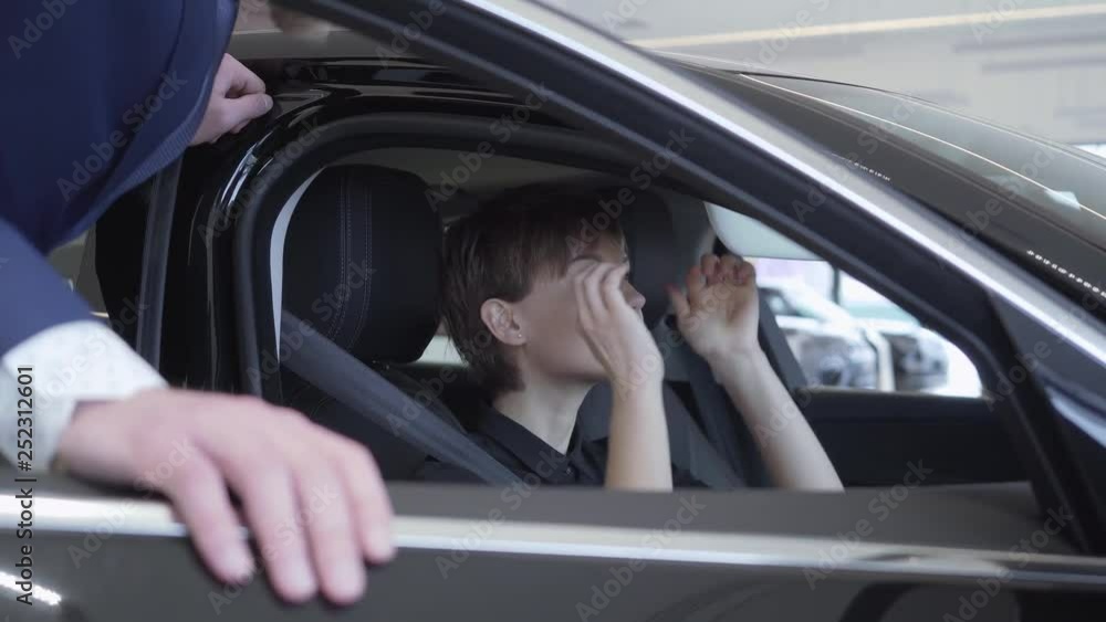 Cute young woman with short hair sits in modern car talking with unrecognizable salesman. Successful woman examines new auto. Professioal salesman in suit consulting customer. Concept of buying Cute young woman with short hair sits in modern car talking with unrecognizable salesman. Successful woman examines new auto. Professioal salesman in suit consulting customer. Concept of buying
