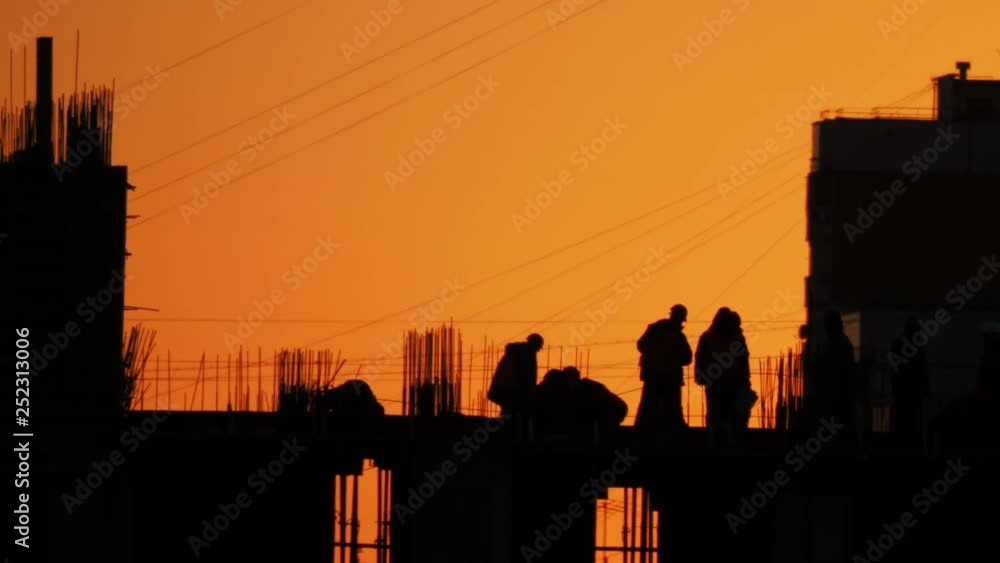 Construction of residential building. Builders go on the unfinished floor with protruding fittings. Silhouettes of workers on the background of an orange sunset. Construction of residential building. Builders go on the unfinished floor with protruding fittings. Silhouettes of workers on the background of an orange sunset.