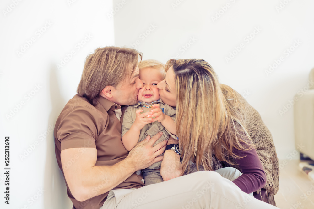 Happy family sitting on floor with their little baby. Family spending time at home with their son. Parents kissing their child.