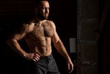 Portrait of topless bearded caucasian Athletic Male Fitness Model posing near dark wall background