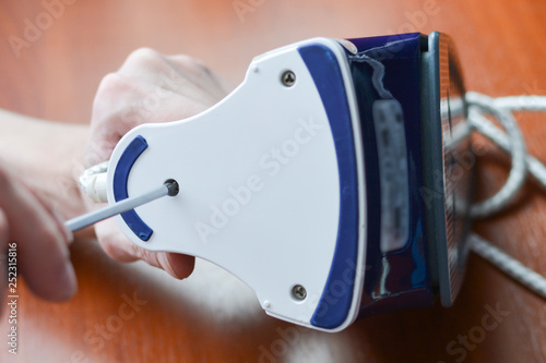 A hand with a screwdriver dismantles the iron. Concept: home appliances repair, maintenance and prevention.