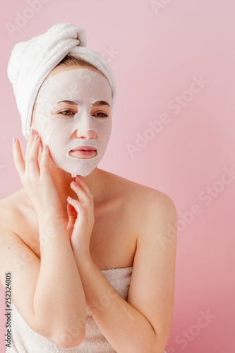 Beautiful young woman is applying a cosmetic tissue mask on a face on a pink background. Healthcare and beauty treatment and technology concept