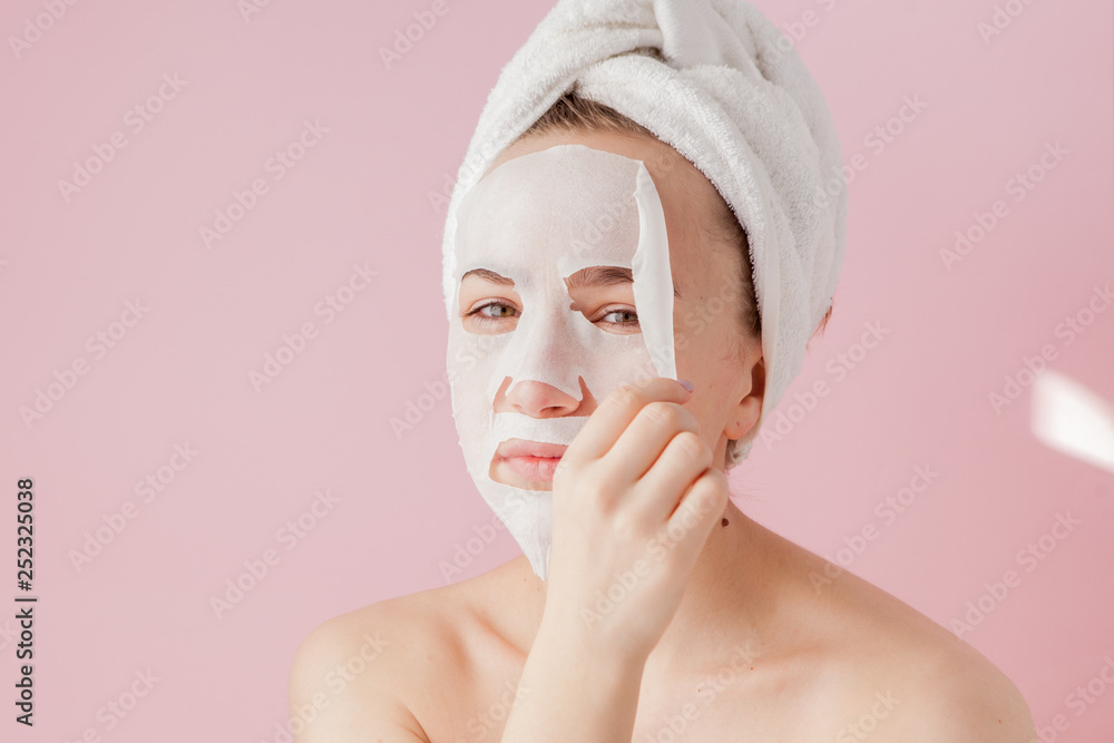 Beautiful young woman is applying a cosmetic tissue mask on a face on a pink background. Healthcare and beauty treatment and technology concept Beautiful young woman is applying a cosmetic tissue mask on a face on a pink background. Healthcare and beauty treatment and technology concept