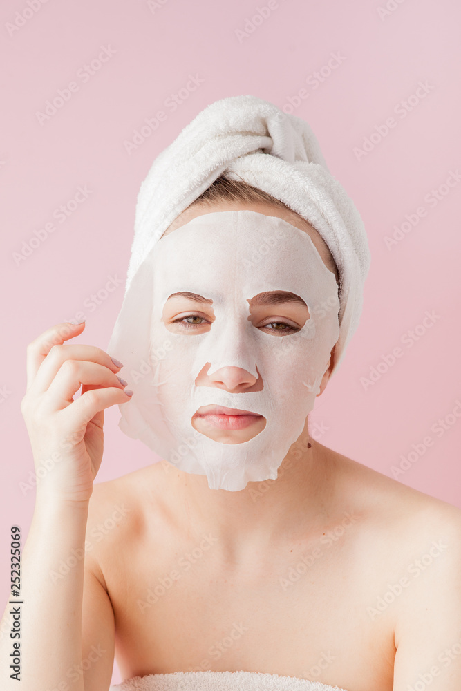 Beautiful young woman is applying a cosmetic tissue mask on a face on a pink background. Healthcare and beauty treatment and technology concept Beautiful young woman is applying a cosmetic tissue mask on a face on a pink background. Healthcare and beauty treatment and technology concept