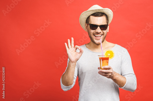 Fototapeta Portrait of attractive young man in hat and sunglasses standing and drinking orange juice over red background