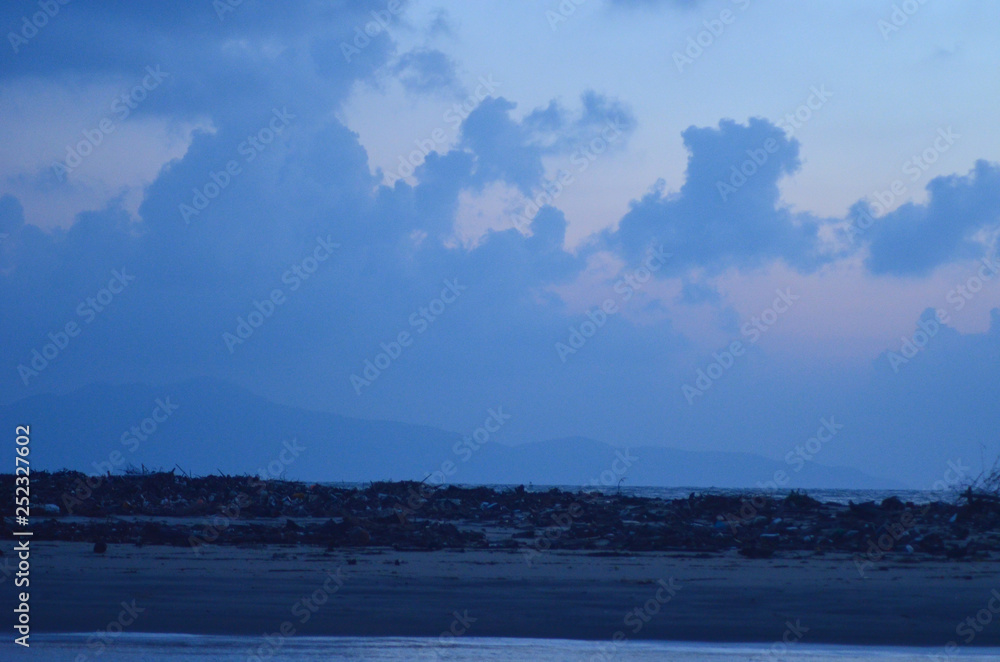 A beach is covered with seaweed and driftwood. Across the bay rise some hills.The sky is filled with clouds. The photograph is in shades of blue, with some pink just touching the sky.