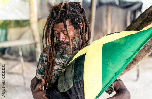 African American bearded male with dreadlocks holding Jamaica flag near tree