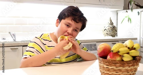 Portrait of a hungry, cute young boy sitting in the kitchen and eating banana at home