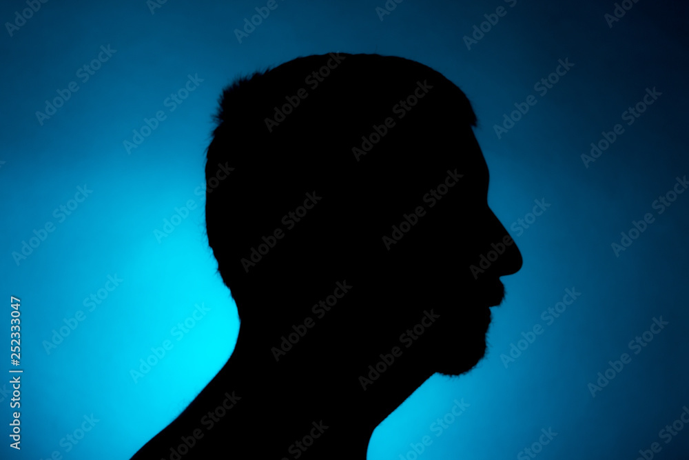 Cleanly defined silhouette of a male person turned to the left against a blue background with a spotlight and bright area right behind the bust. Studio shot with well defined colour background.