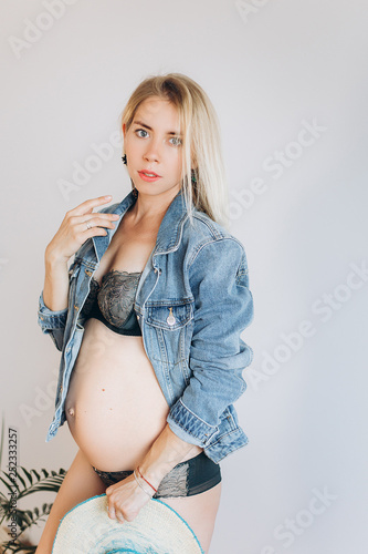 Pregnant cheerful happy future mother expecting for a baby and posing indoors on white background in fashion lingerie and jeans coat with hat. Pregnancy, beauty, fashion concept photo