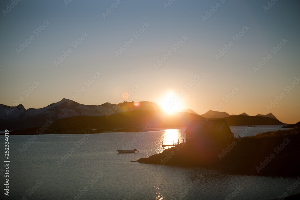 Sunset in a fjord in Norway near Tromsö. Backlit with a few mountains and a silhouette of a house with a boat dock.photographed with wide angle.