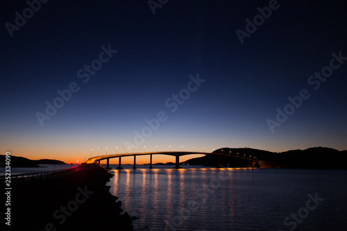 Landscape silhouette and bridge with light on the horizon after sunset. Cloudless, dark blue and symmetrical reflections in the water. Norway nearby Sommaroy. © U_WD