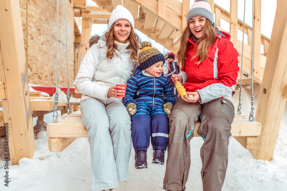 Happy family mom with girlfriend, little son 3-6 years old. Rest in city winter on wooden swing. Happy smiling and having fun. A glass coffee and tea, boiled corn. Winter weekend city park.