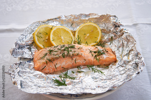 Close up picture salmon fillet baked in aluminium baking foil, low fat, fresh, healty dish served as a lunch or dinner in garden restaurant on white plate and slices of organic lemon and chopped dill.