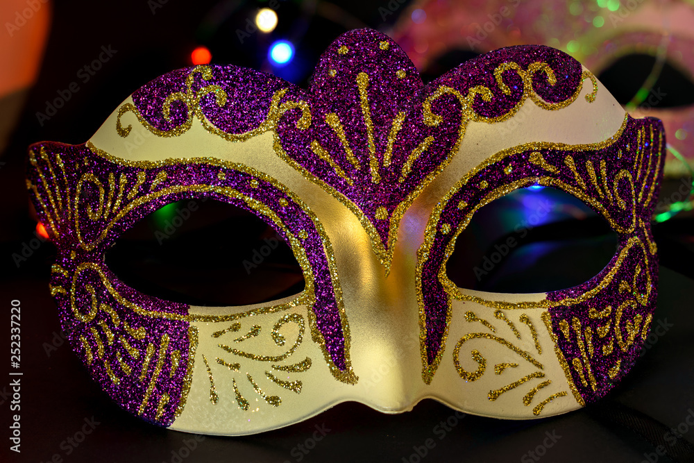 A Venetian mask in purple, gold and yellow with a background of  lights and another mask