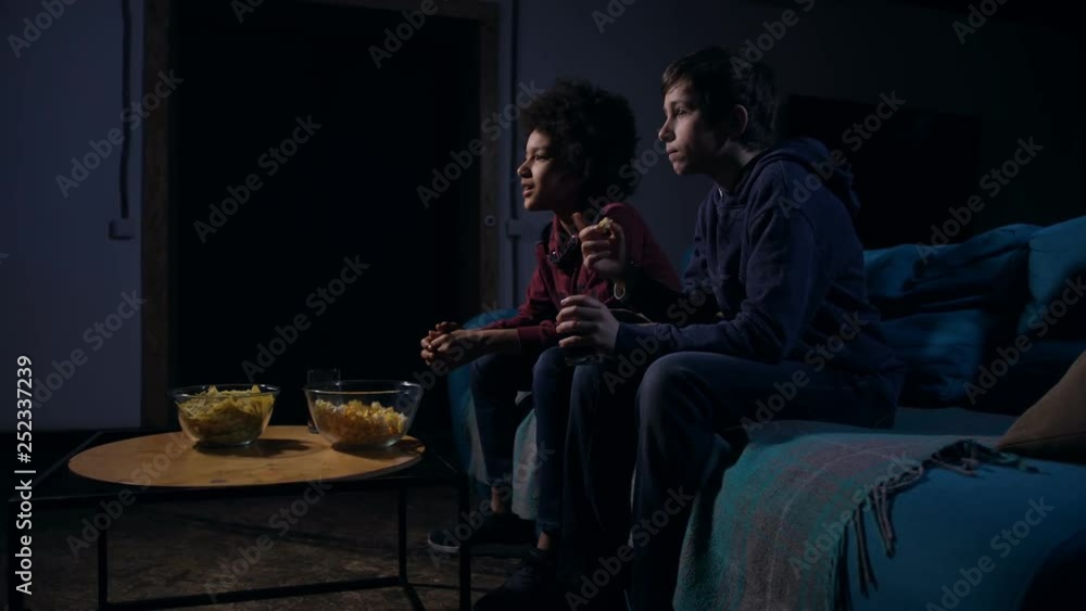 Side view diverse teens slowly taking and putting in mouth popcorn in especially terrible movie scene. Scared friends in casual clothes staring on TV showing horror film sitting on couch in dark room Side view diverse teens slowly taking and putting in mouth popcorn in especially terrible movie scene. Scared friends in casual clothes staring on TV showing horror film sitting on couch in dark room