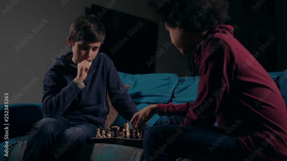 Two focused on game clever teenagers looking at chess board thinking over next move. Diverse concentrated teen friends playing chess sitting on sofa late evening in gloomy domestic room Two focused on game clever teenagers looking at chess board thinking over next move. Diverse concentrated teen friends playing chess sitting on sofa late evening in gloomy domestic room