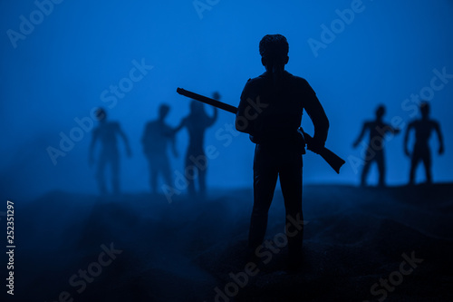 Man with riffle against zombie attack. Zombie apocalypse. Scary view of blurred zombies at cemetery and spooky cloudy sky with fog.