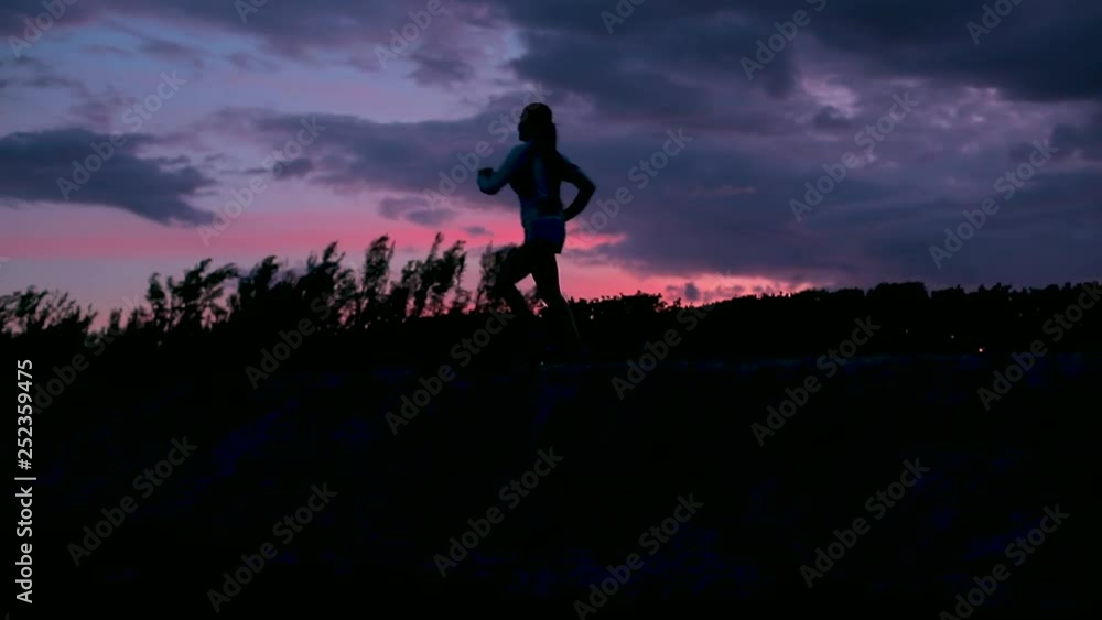 Slow Motion, Silhouette Of A Woman Jogging On A Hill At A Beach With The Sunset Behind Her In Purple, Pink, And Blue Colors Slow Motion, Silhouette Of A Woman Jogging On A Hill At A Beach With The Sunset Behind Her In Purple, Pink, And Blue Colors