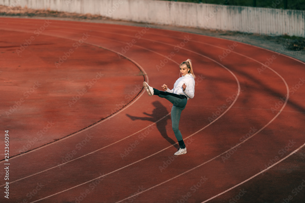 Smiling blonde woman in sportswear kicking in the air while standing on racetrack.
