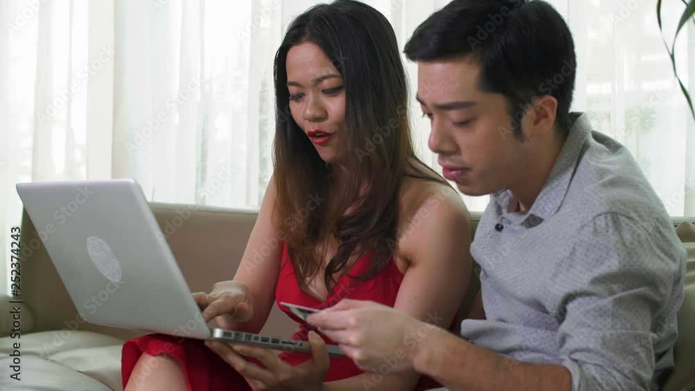 Medium shot of young Asian couple sitting on couch in living room, woman holding laptop, and typing information from credit card which her husband holding in his hand Medium shot of young Asian couple sitting on couch in living room, woman holding laptop, and typing information from credit card which her husband holding in his hand