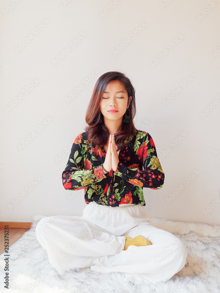 A pretty Chinese young woman practicing yoga at home, sitting on floor with furry cushion with white wall background, exercise, Lotus pose, namaste, working out, Feeling peace and wellness concept. A pretty Chinese young woman practicing yoga at home, sitting on floor with furry cushion with white wall background, exercise, Lotus pose, namaste, working out, Feeling peace and wellness concept.
