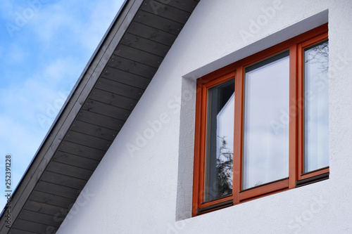 Part of roof construction with grey eaves and brown orange wooden window on white decorative plaster facade with copy space on blue sky. © Inga
