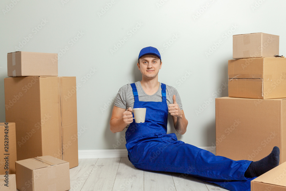 Day moving. Delivery of goods from shopping in the online store. The loader or courier has a break and drinks tea or coffee against the gray wall Day moving. Delivery of goods from shopping in the online store. The loader or courier has a break and drinks tea or coffee against the gray wall