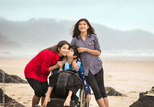 Disabled brother in wheelchair giving older sister kiss on beach