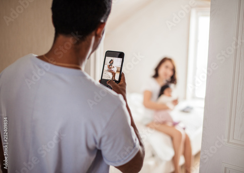 attractiave young mother playing with her baby while daddy taking photos of them at home.blurred background, focus on the photo in the smartphone photo