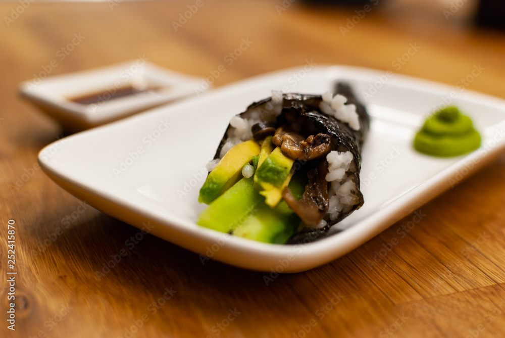 Homemade vegan Temaki-Sushi filled with avocado on a small plate on a wooden table with soy sauce and wasabi