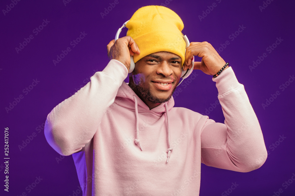 Young African man in hoodie, having the fashion sense of a street urchin, turned a bit in headphones isolated on violet background , holding earphones with hands listening to favorite tracks Young African man in hoodie, having the fashion sense of a street urchin, turned a bit in headphones isolated on violet background , holding earphones with hands listening to favorite tracks