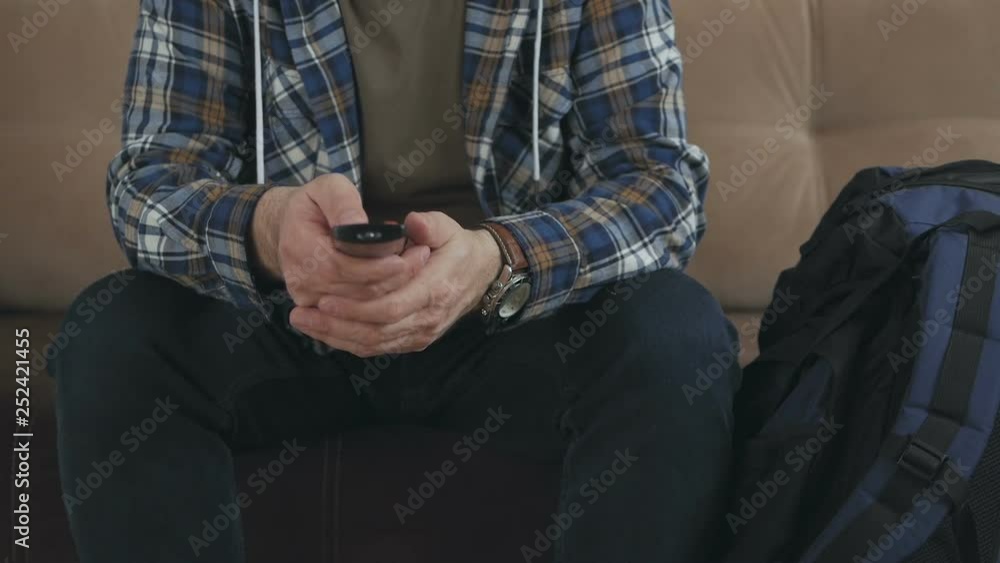A man hipster sitting on the couch presses the buttons of the TV remote control, next to him is a large tourist backpack. A man hipster sitting on the couch presses the buttons of the TV remote control, next to him is a large tourist backpack.