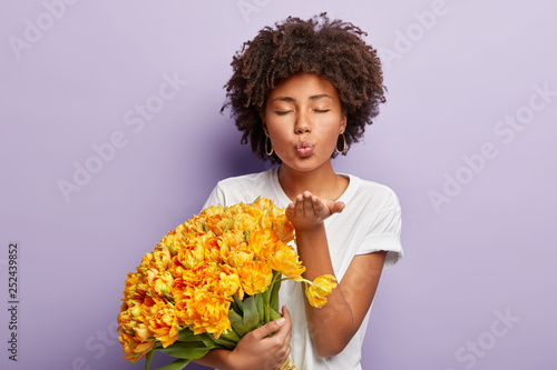 Horizontal shot of lovely affectionate dark skinned woman blows air kiss, spreads palm in front, expresses love and gratitude to husband for beautiful flowers, has eyes shut, isolated over purple wall