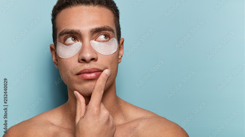 Cropped shot of thoughtful man uses eye patches under eyes, looks upwards, holds chin, stands with bare shoulders, isolated over blue background with blank space aside, reduces wrinkles on face Cropped shot of thoughtful man uses eye patches under eyes, looks upwards, holds chin, stands with bare shoulders, isolated over blue background with blank space aside, reduces wrinkles on face