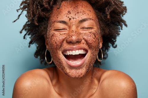 Overjoyed lady with peeling coffee scrub, laughs sincerely, has natural beauty, healthy dark skin, shows white teeth, bare shoulders, cleans face, isolated over blue background. Purity, cosmetology