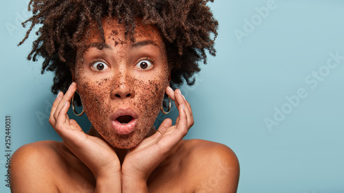 Image of stupefied woman astonished to find out about skin problems, removes pores with coffee scrub, has eyes popped out, tocuhes cheeks, models topless, isolated on blue wall. Wellness concept