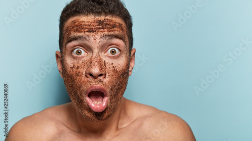 Stupefied emotional young man keeps jaw dropped from amazement, surprised to have spa procedures and notices face with coffee mask in mirror, stands shirtless, isolated over blue background.
