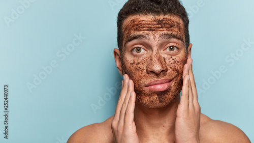 Close up image of curious handsome male touches cheeks, enjoys softness of skin, applies coffee scrub, finds out secrets of beauty from cosmetologist, stands bare shoulders against blue wall