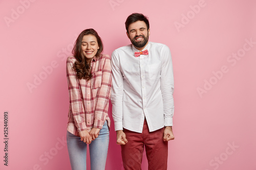Portrait of cheerful female and male clench fists, close eyes from pleasure, satisfied with good results of common work, wear fashionable shirts, isolated over pink background, spend great time