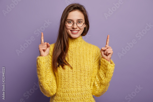 Studio shot of attractive woman advertises something interesting above, wears glasses, yellow jumper, has appealing smile, shows white teeth, isolated over purpe background. Advertising and promotion